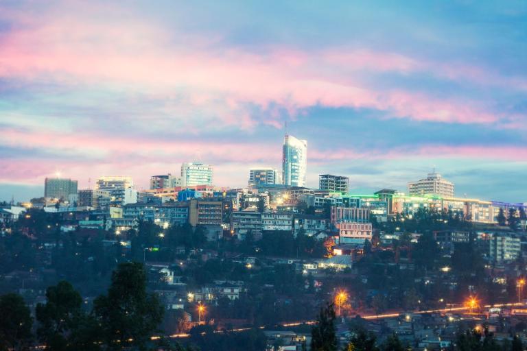 A brightly lit cityscape at dusk with a prominent modern building in the center, surrounded by smaller buildings under a sky with pink and blue hues.