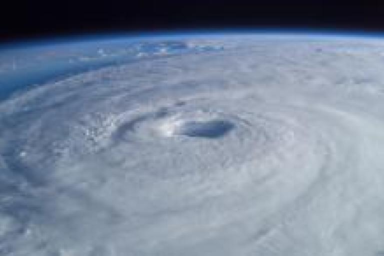 A satellite image of a large, spiraling hurricane with a clearly visible eye at the center, taken from space, showing the swirling cloud patterns over the Earth.