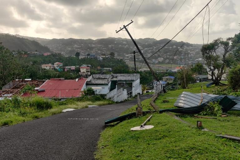 A damaged utility pole leans over a road, with debris and roofing materials scattered around in a hilly residential area. Houses and trees are visible in the background under a cloudy sky.
