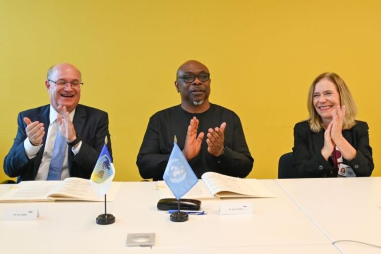 Three people sit at a table with documents and small flags in front of them, clapping their hands with smiles. The background is a yellow wall.