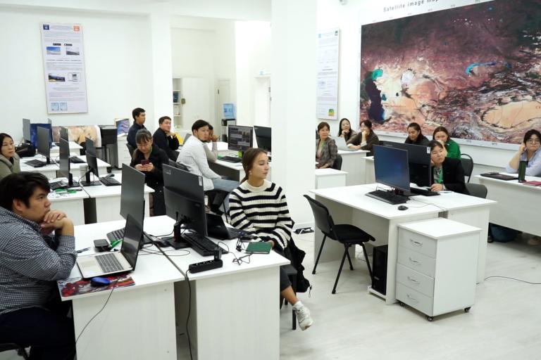 A classroom with several students sitting at desks with computers. They are attentively facing the front where an instructor is visible. A large, detailed map is displayed on the wall.