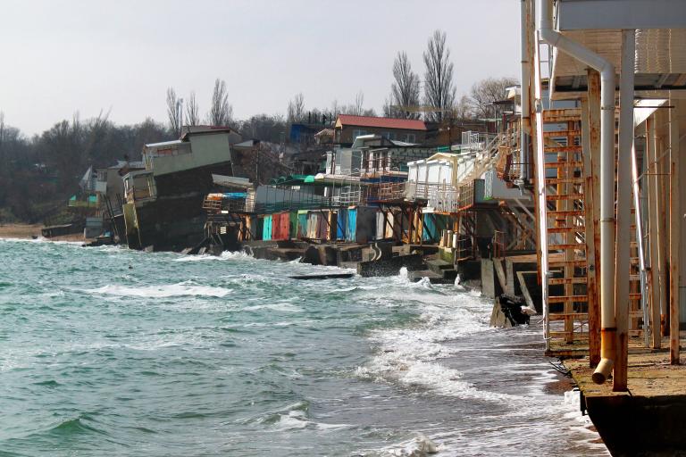 Beachfront buildings extensively damaged by erosion with portions collapsed into the sea, and waves lapping against the remains of the structures. Trees are visible in the background.