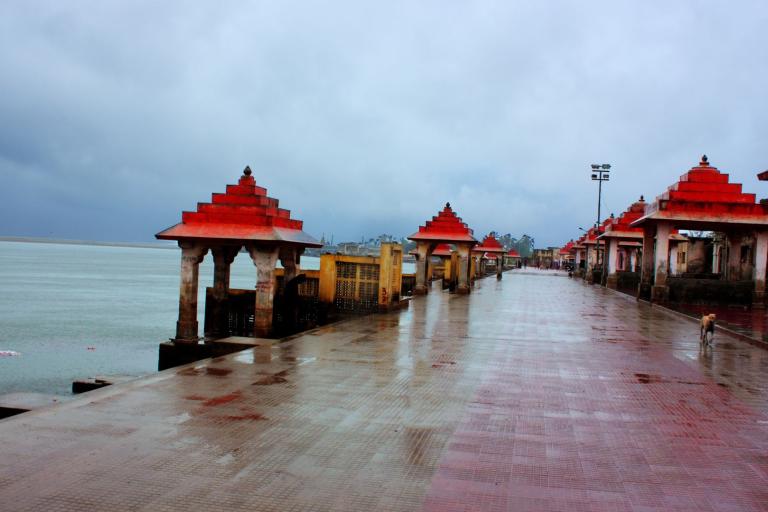 A riverside promenade with red-roofed pavilions on a rainy day. The ground is wet, and the sky is overcast. The walkway is empty except for a lone dog.