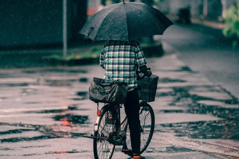 Person riding a bicycle in the rain, holding a large black umbrella and carrying two bags on the bike. The street is wet with visible puddles.