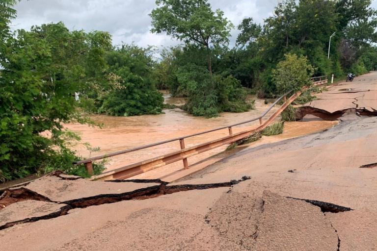 A damaged road with severe cracks and a collapsed section due to flooding; muddy water flows beneath, surrounded by trees.