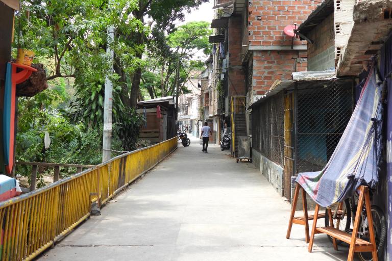 Narrow alley with brick buildings on one side, trees on the other, a yellow railing, and a person walking away in the distance.