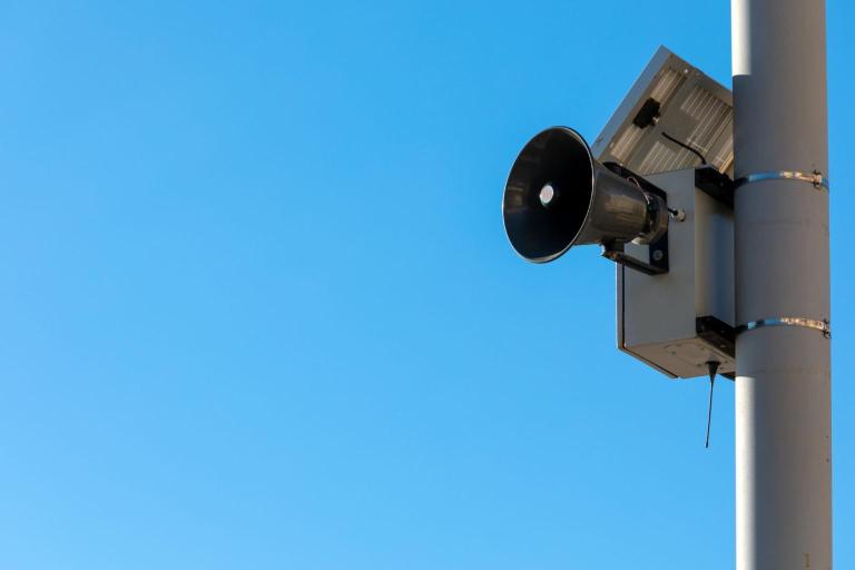 Outdoor speaker mounted on a pole against a clear blue sky.