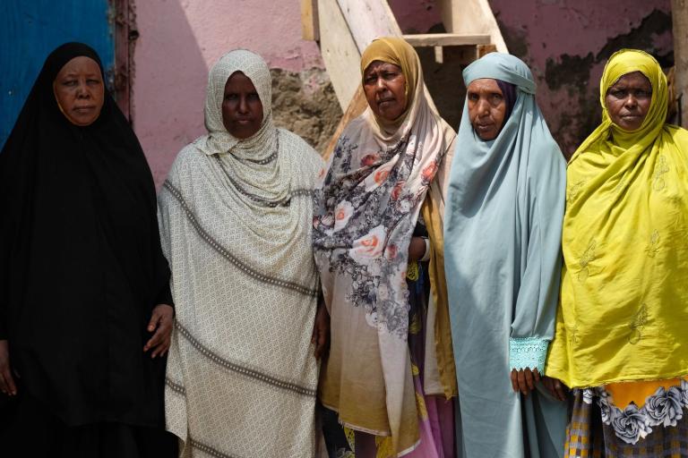 Five women stand side by side, wearing various colored hijabs. The background features a pink wall and a staircase.