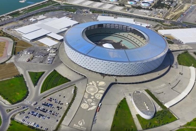 Aerial view of a large, modern stadium with a circular roof, surrounded by parking areas and greenery, located near a body of water.