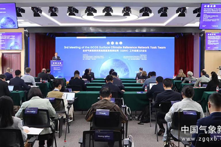 A conference room with attendees viewing a large screen displaying the "3rd Meeting of the GCOS Surface Climate Reference Network Task Team." Chairs and tables are arranged in rows.