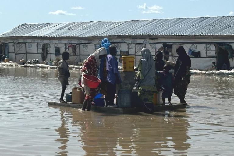 People gather with containers on a raft in floodwater near a building with a metal roof.