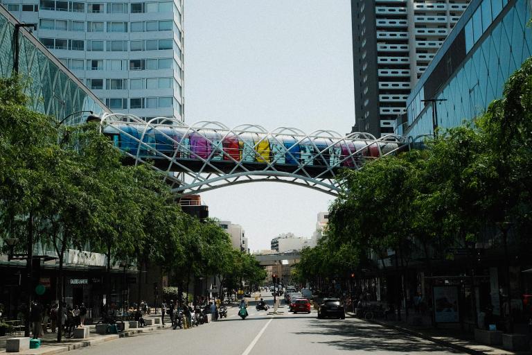 A street view showing a colorful pedestrian bridge between two tall buildings, with trees lining the road and vehicles in the distance.