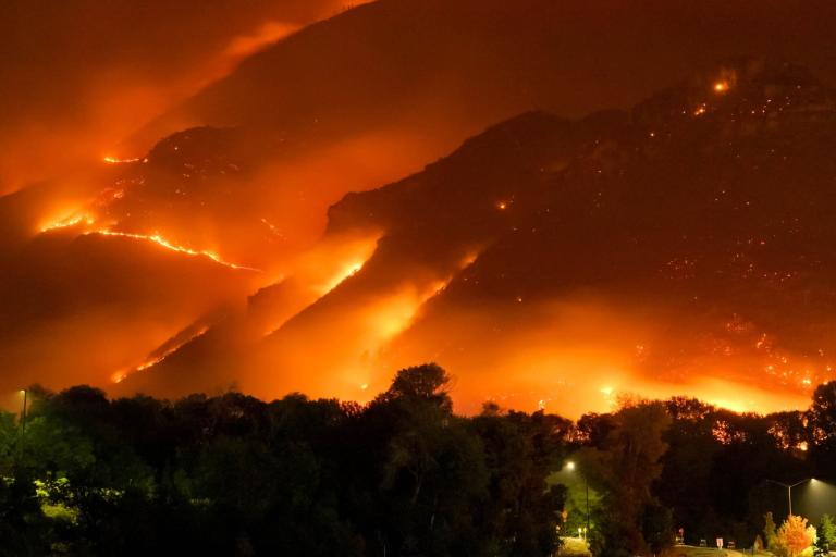 A wildfire engulfs a mountainside at night, with intense orange flames and smoke illuminating the scene. Trees and a few lights are visible in the foreground.