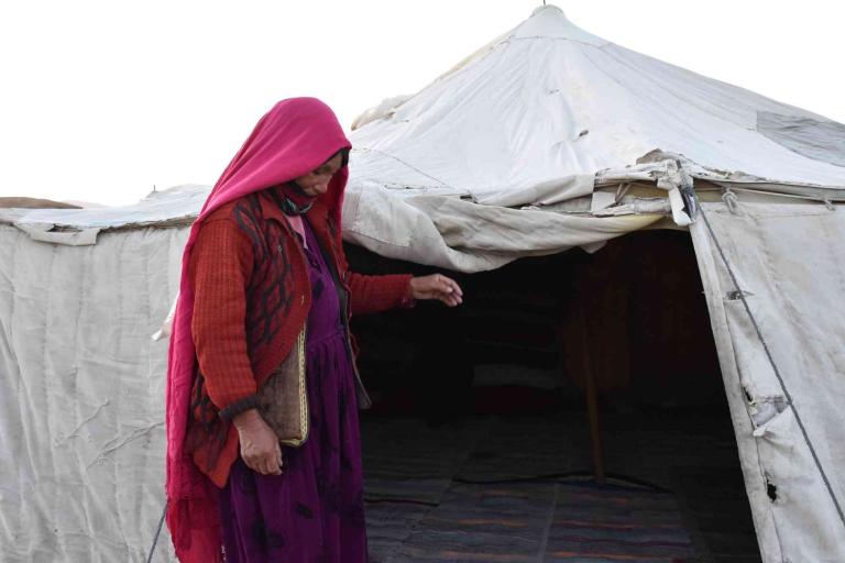 A person wearing a pink headscarf and red sweater stands outside a white tent.