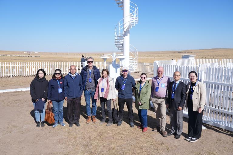 A group of people standing together outside near a spiral staircase and a white fence in a field under a clear blue sky.