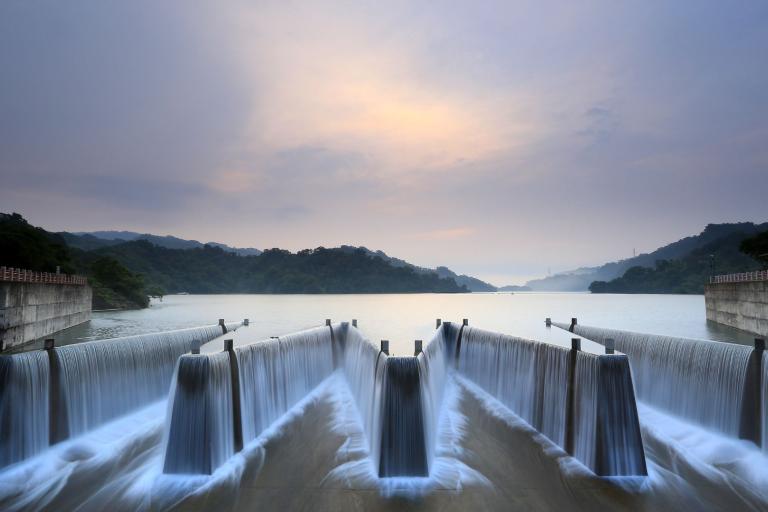 A symmetrical dam with flowing waterfalls under a cloudy sky, surrounded by lush green hills and a tranquil lake.