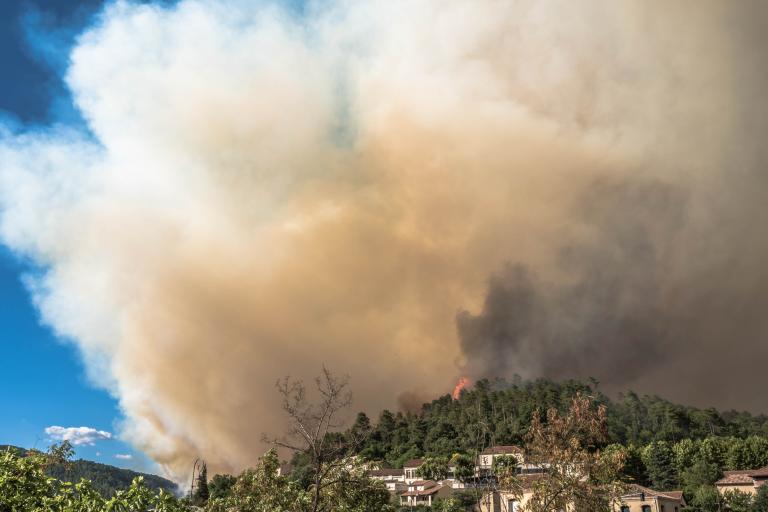 Thick smoke billows from a forest fire near a town, with red flames visible among the trees under a clear blue sky.