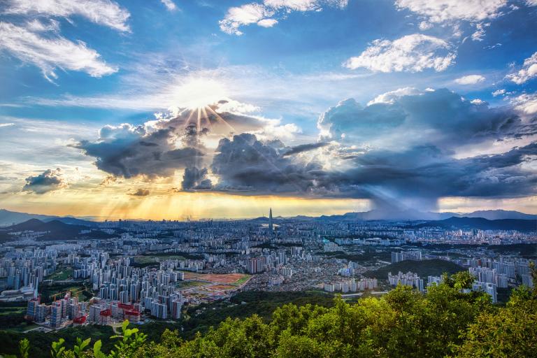 Panoramic view of a cityscape with dramatic clouds and sun rays breaking through. A rainstorm is visible in the distance, surrounded by green hills and urban buildings under a blue sky.