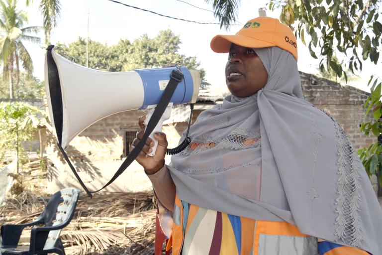 A woman in a gray headscarf and orange cap speaks into a megaphone outdoors, near a tree and some debris.