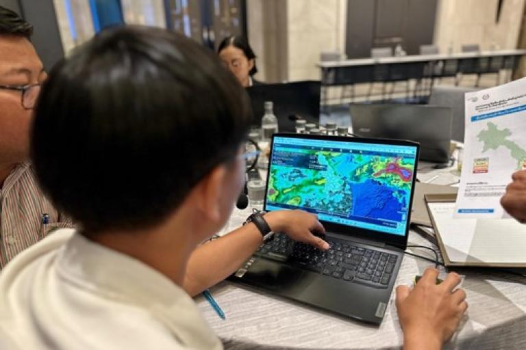 Person studying a colorful map on a laptop screen in an office setting, with papers and colleagues nearby.