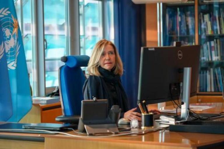 A person sitting at a desk, working on a computer in an office. A United Nations flag and bookshelf are visible in the background.