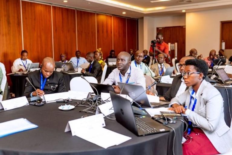 A group of people attending a conference in a meeting room, seated at round tables with laptops and notepads.