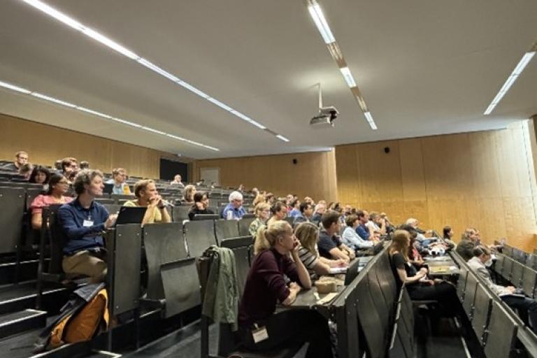 A lecture hall with attendees seated in rows, facing the front. The room has wooden walls and overhead lighting.