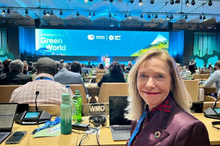 A woman in a conference hall sits at a desk with various electronic devices. A large screen displays "In Solidarity for a Green World" in the background.