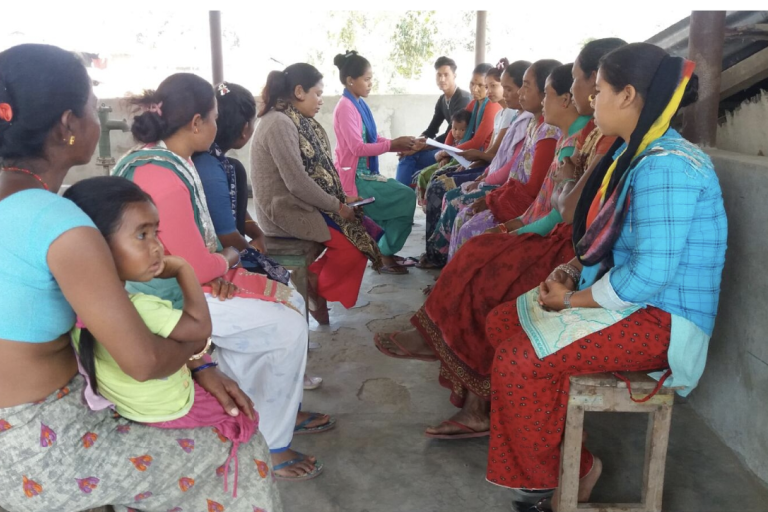 A group of people sit in a circle outdoors, some holding notebooks, engaged in discussion. A child is seated on one person's lap.