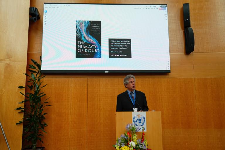 A man stands at a podium giving a presentation in front of a large screen displaying the book cover of "The Primacy of Doubt" by Tim Palmer.
