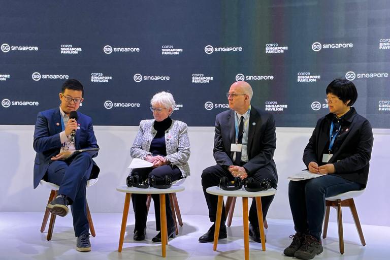 Four individuals are seated on stage, engaging in a panel discussion. Behind them is a backdrop with logos and text. Microphones and headphones are placed on the small tables in front of them.