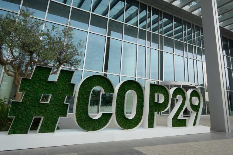Large green hashtag sign reading "#COP29" outside a modern glass building with trees nearby.