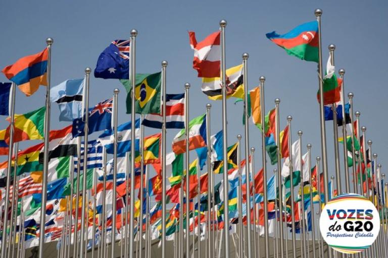 Numerous international flags on poles, waving against a clear sky. A logo in the corner reads "Vozes do G20 Perspectivas Cidadãs.