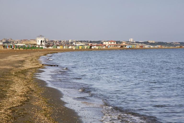A sandy beach with gentle waves, bordered by a town with buildings and colorful beach huts in the background under a clear sky.