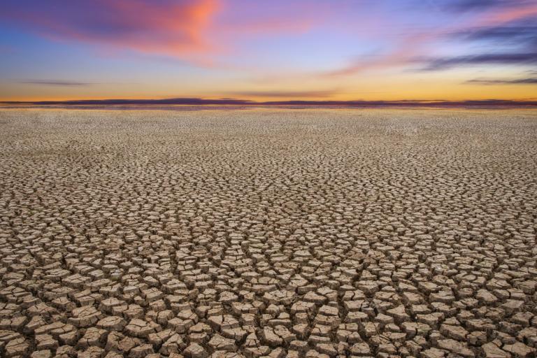 Cracked dry earth stretches toward a colorful sunset sky with pink and orange clouds on the horizon.