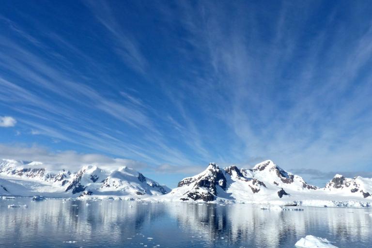 Snow-covered mountains and glaciers are reflected in calm Antarctic waters under a blue sky with wispy clouds.