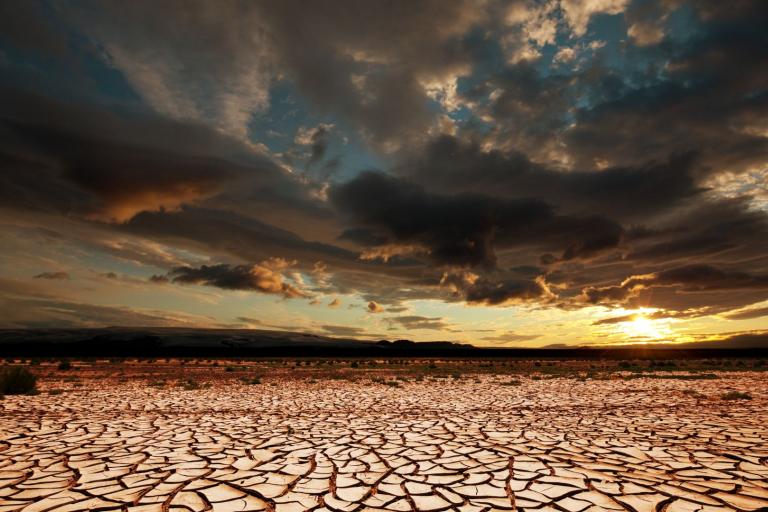 A cracked, dry desert landscape with dramatic clouds overhead and the sun setting on the horizon.