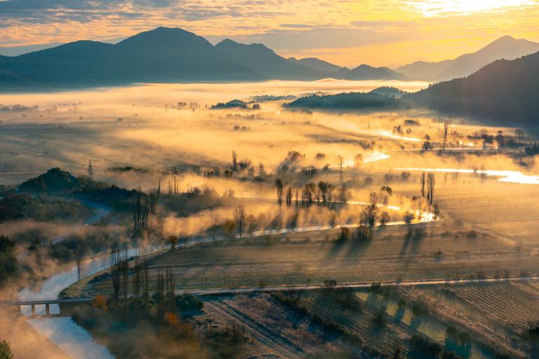 Aerial view of a misty landscape featuring meandering rivers, fields, and distant mountains at sunrise.