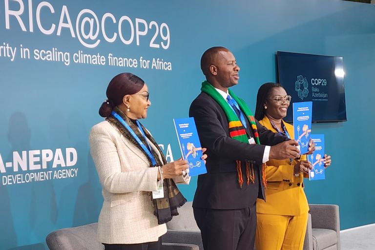 Three people stand holding booklets in front of a backdrop reading "AFRICA@COP29: Solidarity in scaling climate finance for Africa," featuring AUDA-NEPAD and COP29 branding.