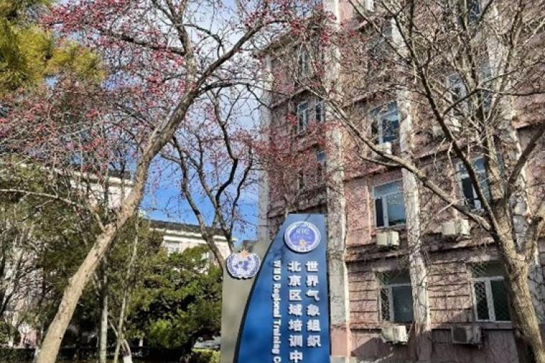 A blue sign with white text stands in front of a building with pink blossoms and trees. The building has multiple windows and an air conditioning unit.