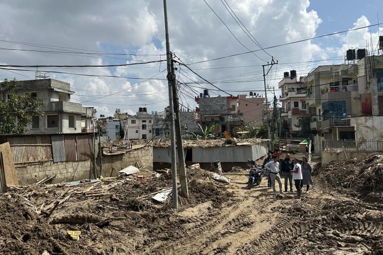 A group of people walk through a muddy, debris-filled street in a residential area, with damaged buildings and clouds in the sky.