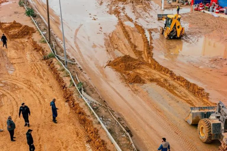 Flooded street with muddy water, people walking, and construction vehicles working near a PetroOil station.