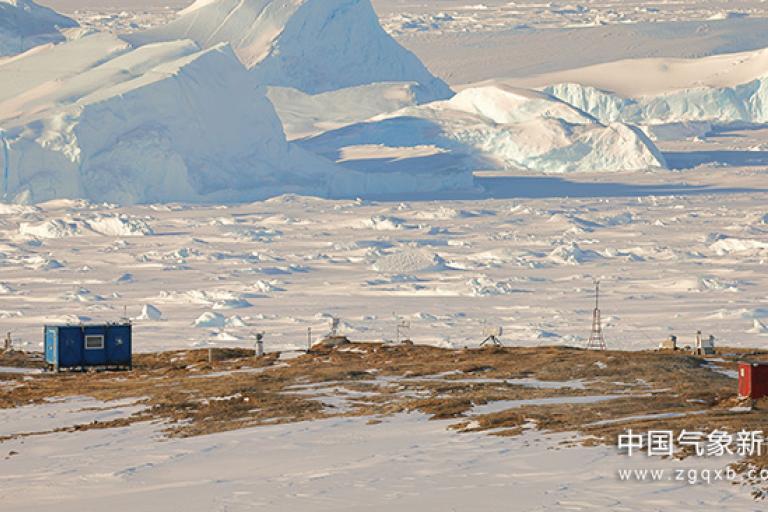 Two small structures on a barren landscape with snow and large ice formations in the background.