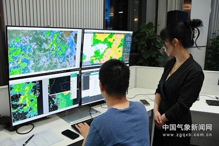 Two people in an office view weather maps and data on multiple monitors in a control room setting.