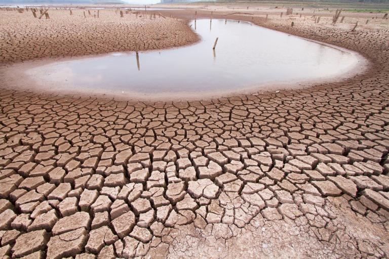 Cracked, dry earth surrounds a small pool of water in an arid landscape under a cloudy sky.