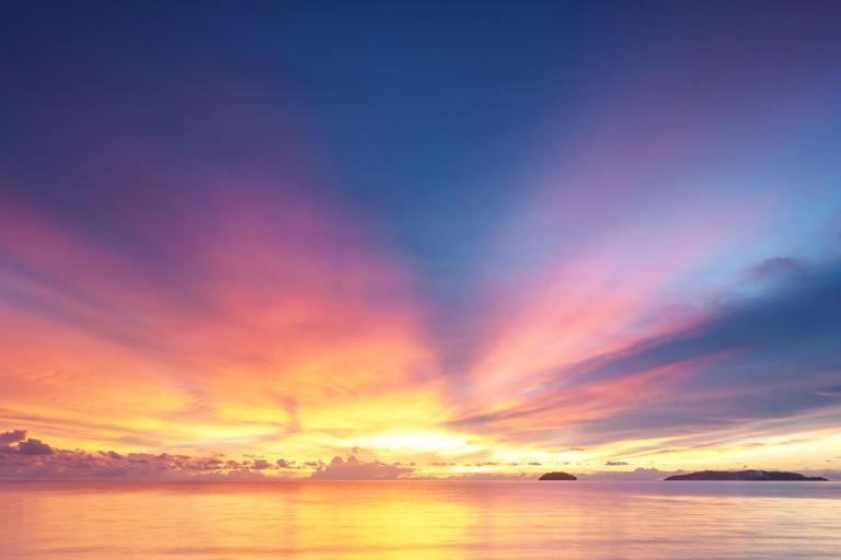 A vibrant sunset over calm waters, with a sky transitioning from orange and yellow near the horizon to deep blue above, and silhouetted islands on the right.