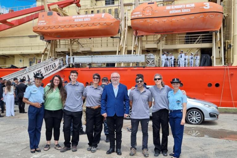 A group of people stands in front of a red ship with lifeboats. The ship's name includes "Irizar" and "Buenos Aires." Some individuals wear uniforms, and a car is visible nearby.