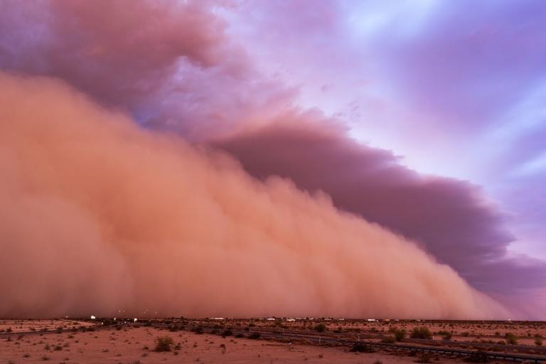A large dust storm moves across a desert landscape beneath a colorful sky.