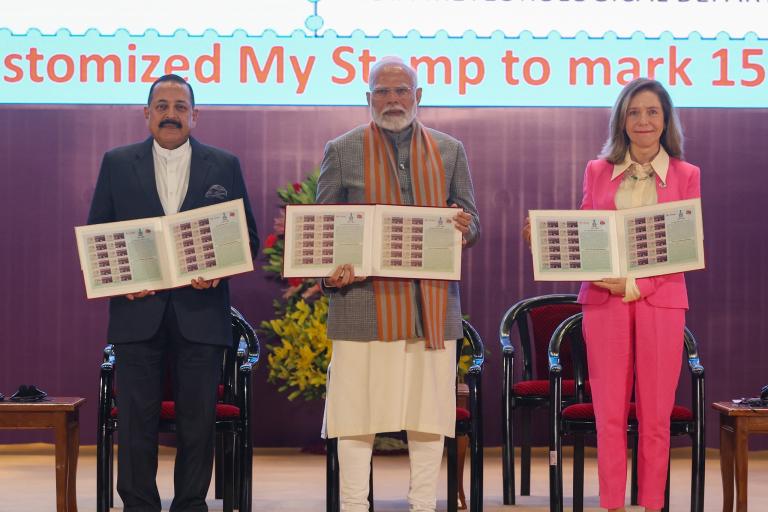 Three individuals stand holding stamp sheets at a ceremony. A sign in the background mentions "India Meteorological Department" and "Customized My Stamp.