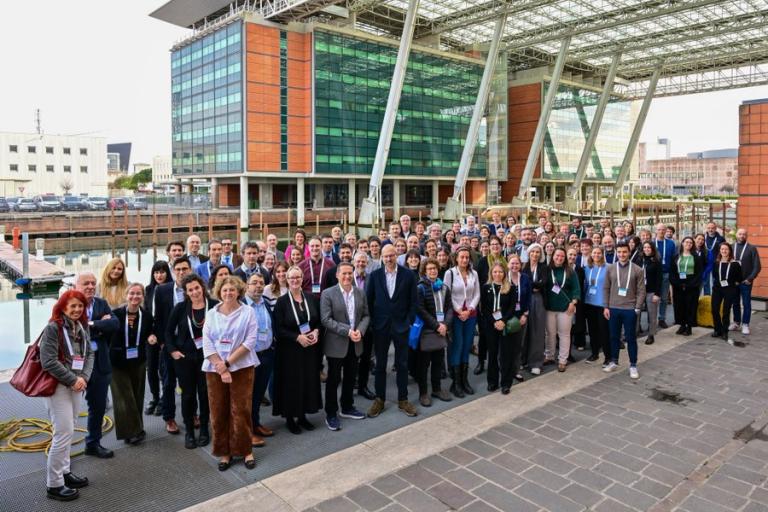A large group of people pose for a photo outside a modern building with glass and brick elements. They stand on a paved area, some wearing lanyards. The setting appears to be a professional event.